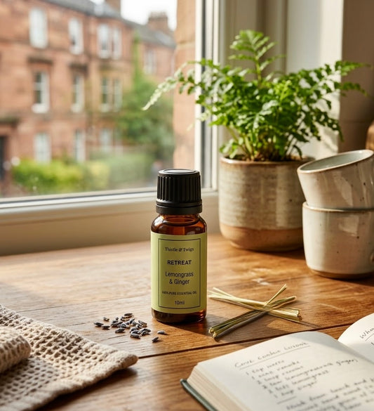 Thistle & Twigs Retreat Lemongrass & Ginger essential oil, 10ml amber bottle on a sunlit wooden table beside a potted fern, ceramic cups and an open notebook.