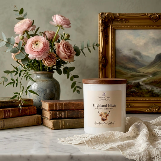Highland Elixir Soy Wax Natural Candle by Thistle & Twigs on a marble surface with pink ranunculus, antique Scottish books and a Highland landscape painting in a warm elegant setting