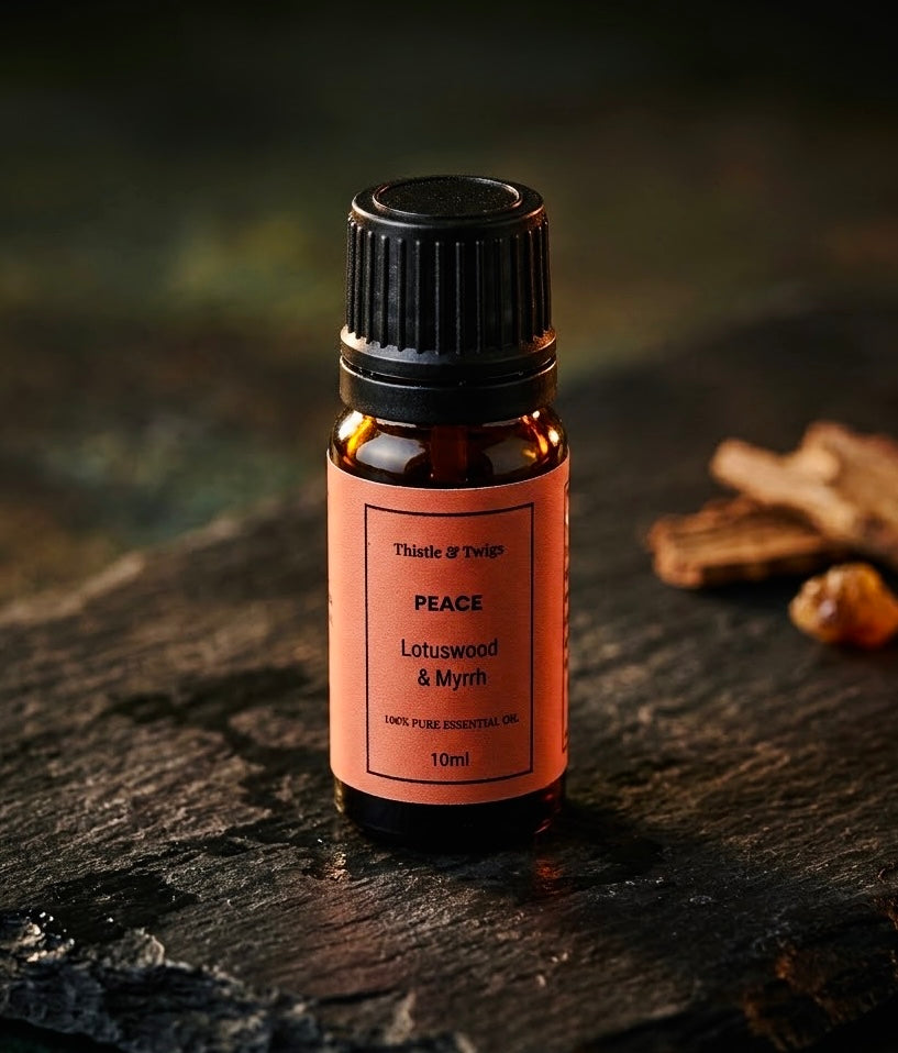 Thistle & Twigs Peace Lotuswood & Myrrh essential oil, 10ml amber bottle with a coral label and black cap, displayed on rustic dark wood beside wood and resin pieces.