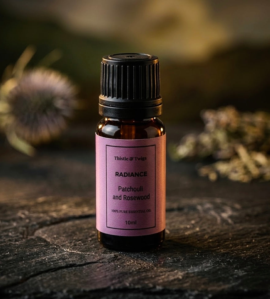 Thistle & Twigs Radiance Patchouli and Rosewood essential oil, 10ml amber bottle with a pink label and black cap, displayed on rustic dark wood with dried botanicals in the background.