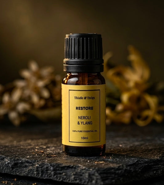 Thistle & Twigs Restore Neroli & Ylang essential oil, 10ml amber bottle with a yellow label and black cap, displayed on a rustic dark surface with flowers in the background.
