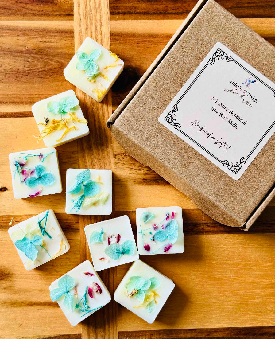 Thistle & Twigs 9 Luxury Botanical Soy Wax Melts with pressed blue hydrangeas and pink petals scattered around a kraft gift box on a warm wooden surface, hand poured in Scotland