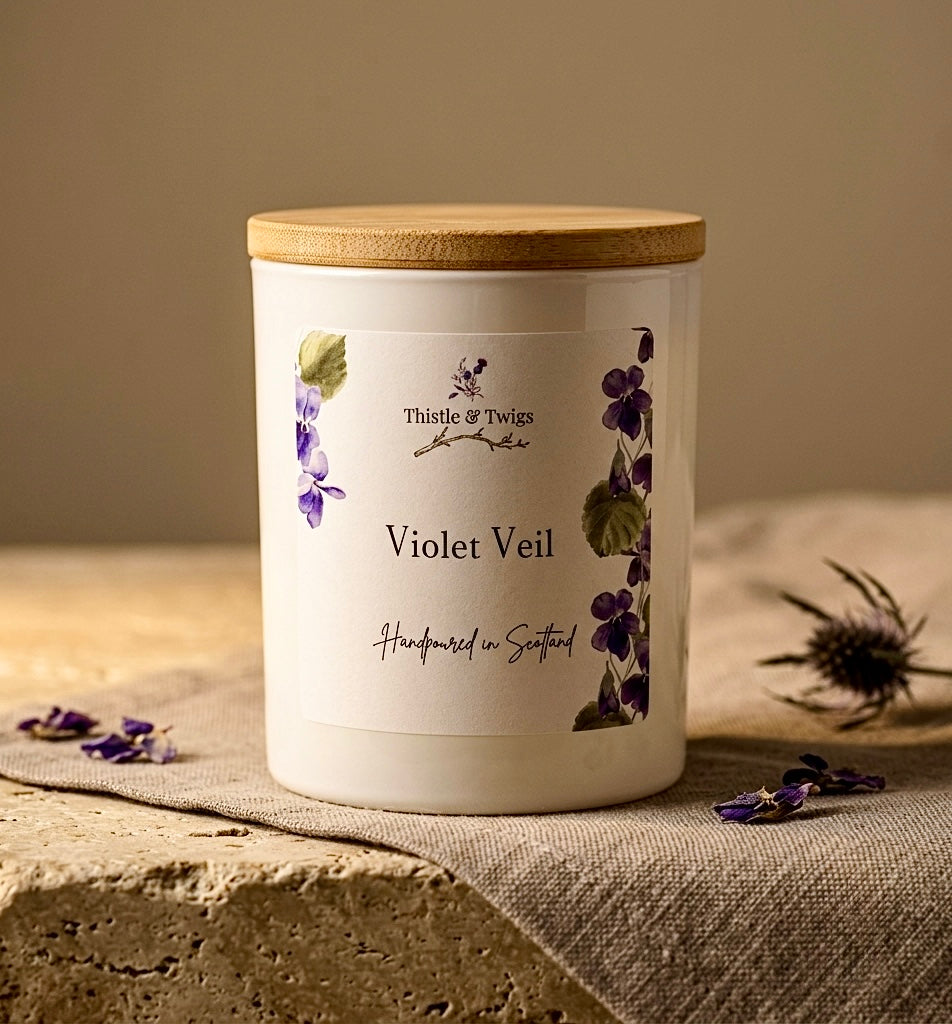 Thistle & Twigs Violet Veil handpoured soy wax candle with bamboo lid and botanical violet label, displayed on a stone surface with linen cloth, dried violet petals and Scottish thistle