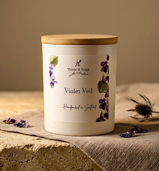Thistle & Twigs Violet Veil handpoured soy wax candle with bamboo lid and botanical violet label, displayed on a stone surface with linen cloth, dried violet petals and Scottish thistle